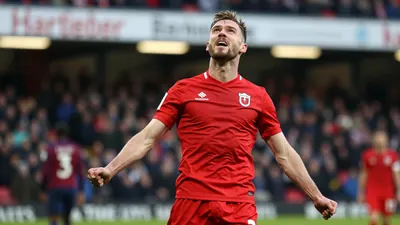 A Hartlepool striker in a red shirt with sleeves rolled up, mid-celebration after scoring a header, face lit by floodlights