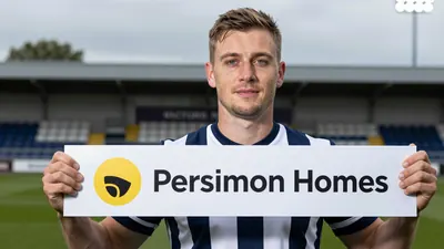 A Hartlepool United staff member in a navy blue and white striped shirt stands at Victoria Park, holding a Persimmon...