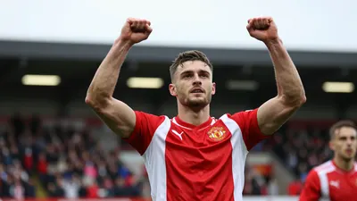 A Hartlepool United player in a red and white striped jersey, mid-20s, muscular build, celebrating a goal with arms raised