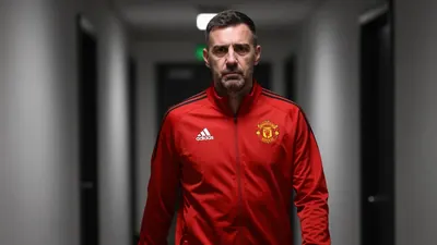 A 35-year-old male assistant coach in a Hartlepool United tracksuit, with a serious expression, standing in a dimly lit...